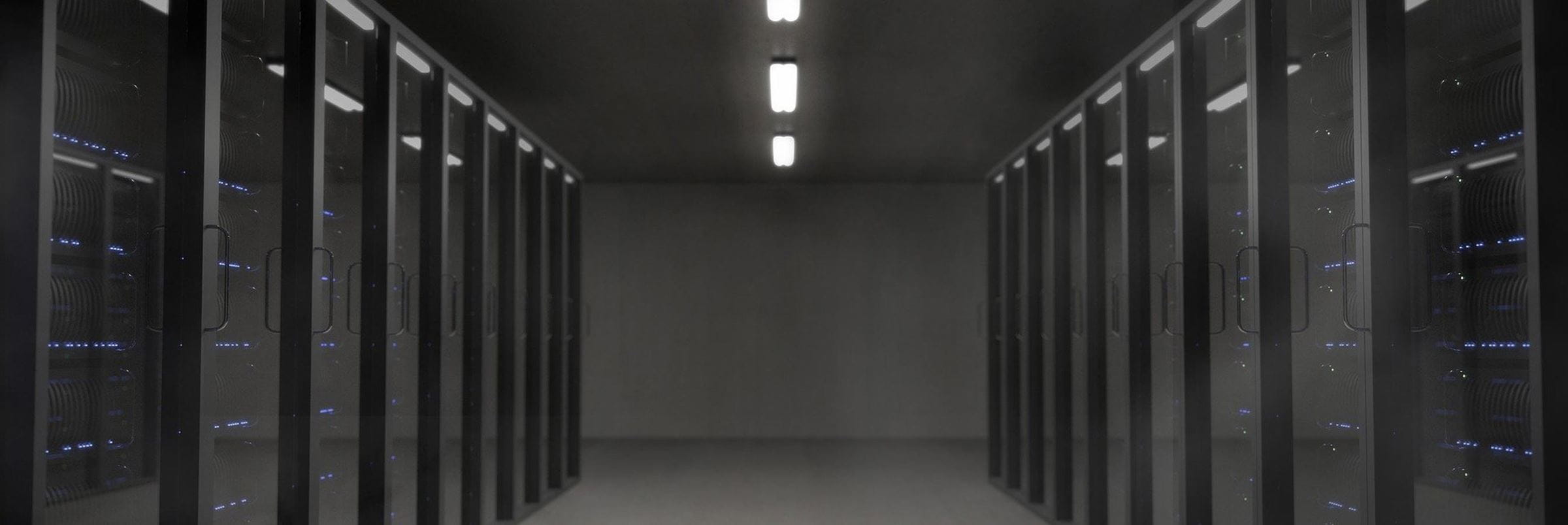 Two rows of data centre server racks in black, with a central path. A central strip of lights overhead providing sparse light to the gloomy conditions.
