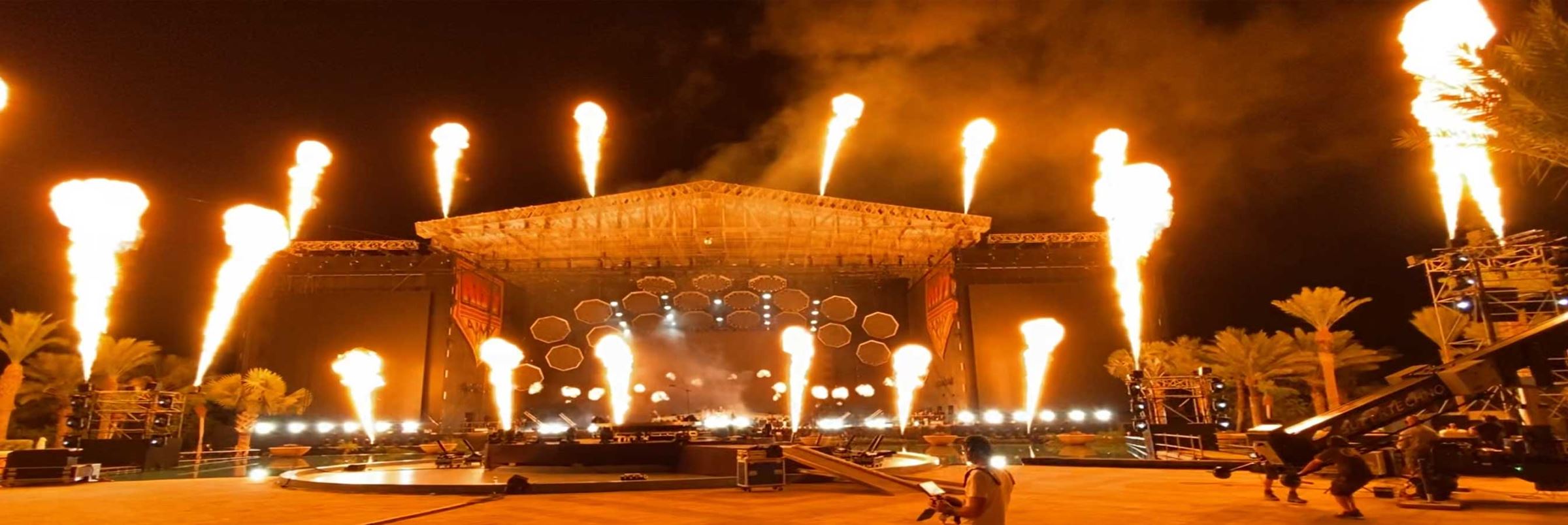 A massive stage is dotted with daunting flames and a mass of lights scattered al around. The stage also has a massive screen.