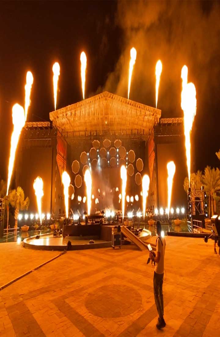 A massive stage is dotted with daunting flames and a mass of lights scattered al around. The stage also has a massive screen.
